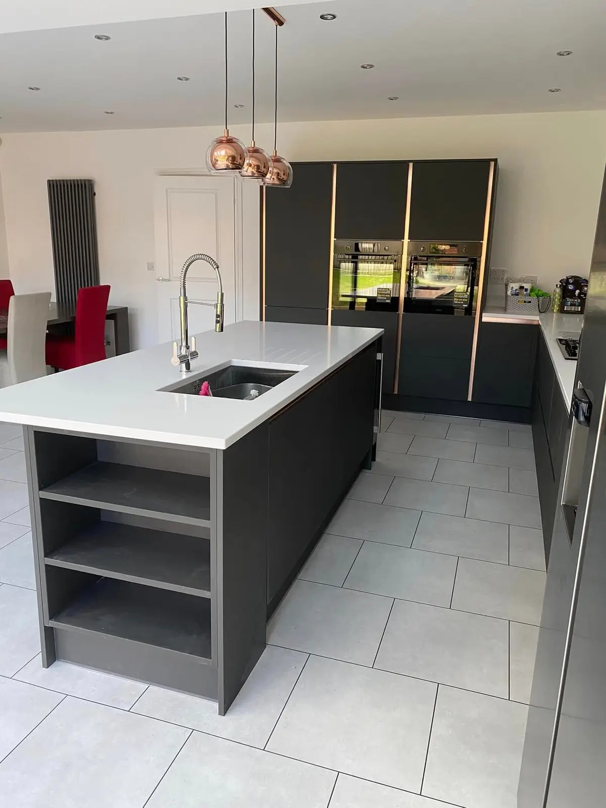 Kitchen island with open-end shelving, white worktop and undermount sink, in front of tall dark grey units with copper profiles
