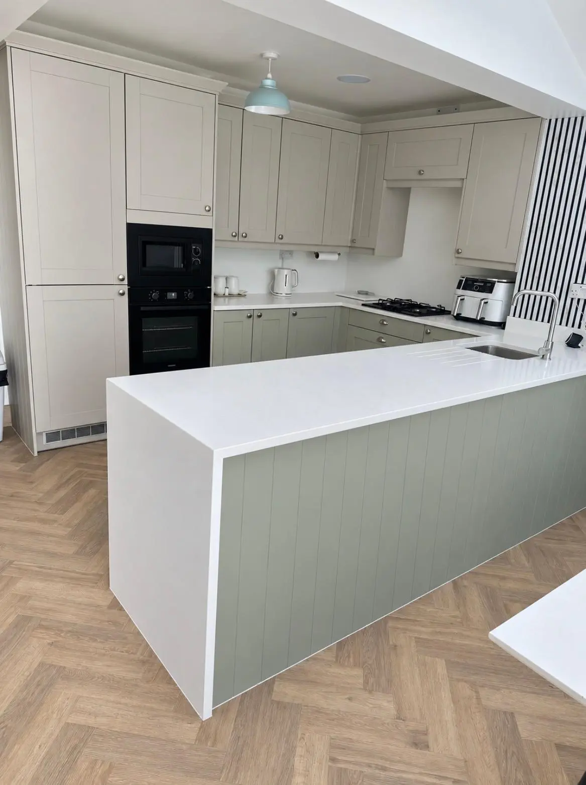 Green sage shaker kitchen with cashmere wall units, a white quartz peninsula with waterfall edge and built-in black ovens on a herringbone floor