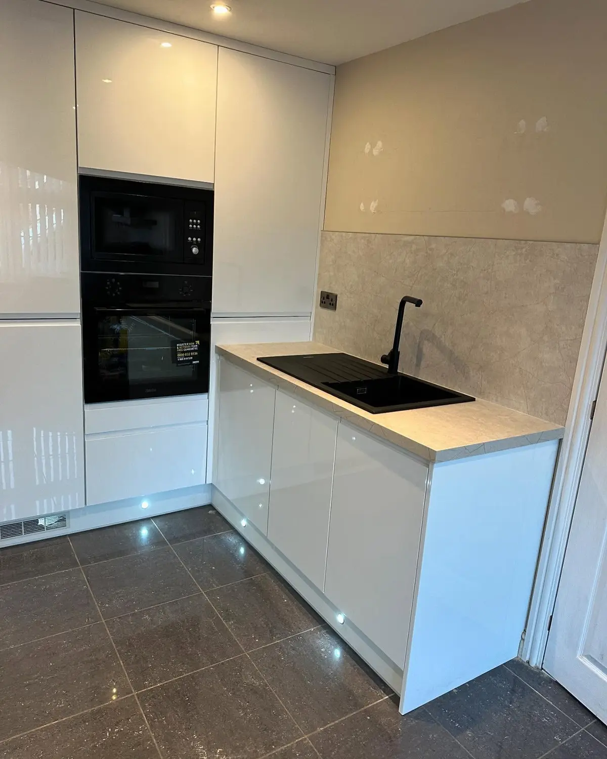 White high-gloss handleless kitchen with tall housing, black built-in ovens, stone-effect worktop and plinth lights